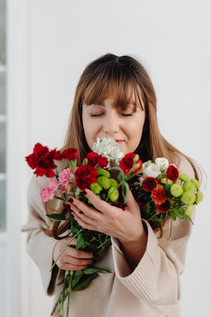 A woman with long brown hair and closed eyes holds a vibrant bouquet of fresh flowers close to her face, appearing to enjoy their scent. The arrangement includes red carnations, pink roses, white hydr