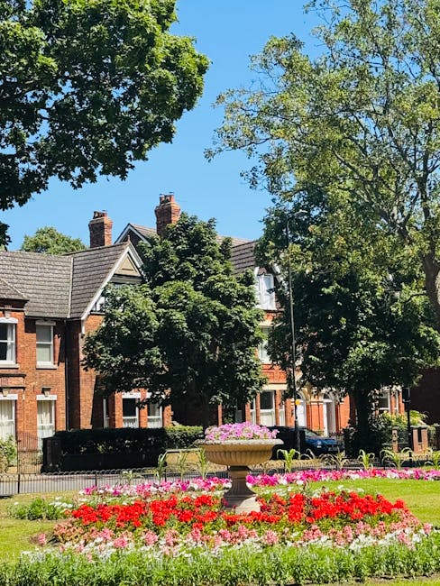 A view of a lush, green park area with vibrant flower beds in a variety of red, pink, and white blooms, arranged in a neat, circular pattern around a large stone planter filled with pink flowers. Tall