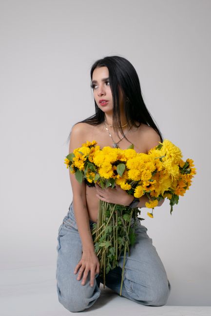 A young woman with long black hair and light skin kneels on the floor against a plain light gray background, holding a large bouquet of bright yellow chrysanthemums with green stems and leaves. She is