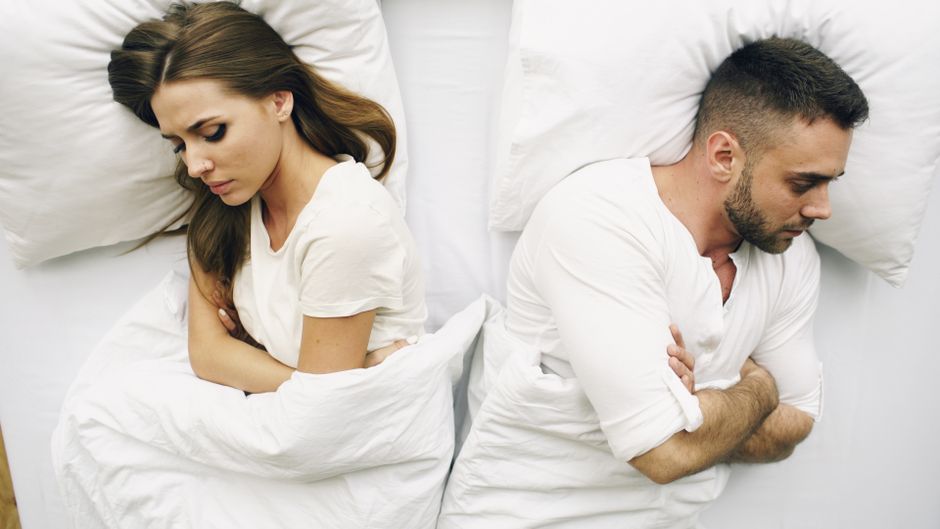 A woman and a man are lying on separate white pillows in a bed with white bedding, positioned back-to-back and facing away from each other with their arms crossed. The woman has long brown hair, and h