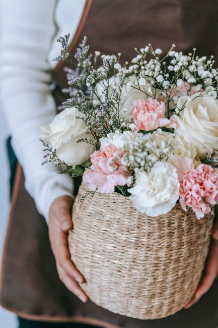 A person holding a woven wicker basket filled with a floral arrangement featuring white roses, pink carnations, and sprigs of baby's breath and lavender. The flowers appear fresh, with delicate petals