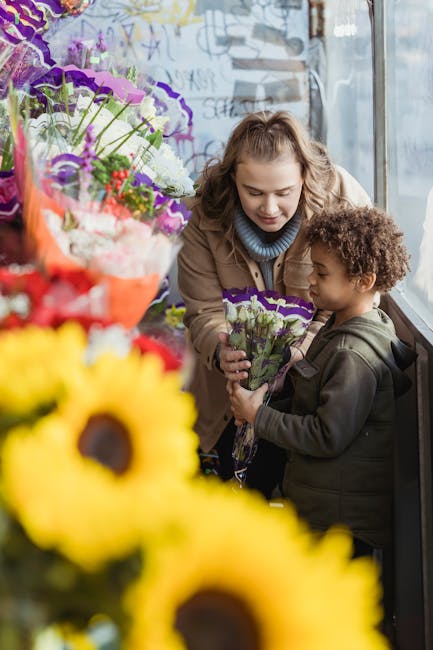 A young woman with wavy blonde hair, wearing a beige jacket and a blue scarf, is smiling as she presents a bouquet of purple and white fresh flowers to a small boy with curly brown hair, dressed in a 